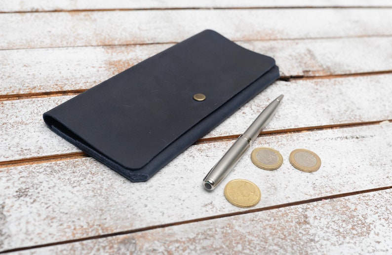 May include: A dark blue leather wallet with a silver pen and three gold coins on a white wooden surface.