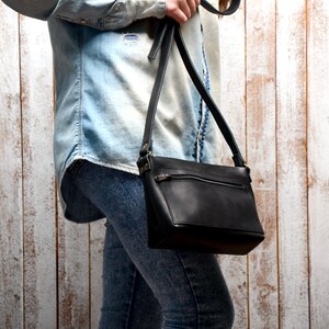 Peut inclure: Un sac &agrave; bandouli&egrave;re en cuir noir avec une poche zipp&eacute;e et une longue bandouli&egrave;re r&eacute;glable. Le sac est port&eacute; par une personne v&ecirc;tue d'une chemise en jean bleu clair et d'un jean.