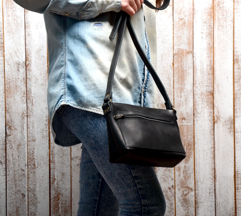 Peut inclure: Un sac &agrave; bandouli&egrave;re en cuir noir avec une poche zipp&eacute;e et une longue bandouli&egrave;re r&eacute;glable. Le sac est port&eacute; par une personne v&ecirc;tue d'une chemise en jean bleu clair et d'un jean.