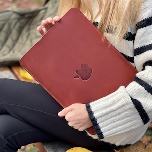 May include: A reddish-brown leather laptop sleeve with the Apple logo cutout. The sleeve is held by a person wearing a black and white striped sweater. The sleeve appears to be new and unused.