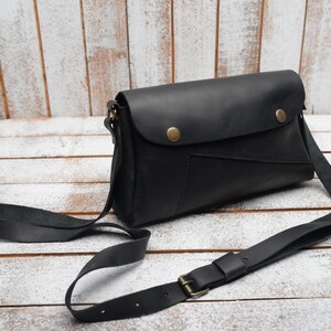 May include: A black leather crossbody bag with a flap closure and a long adjustable strap. The bag has a small pocket on the front and two brass snaps.