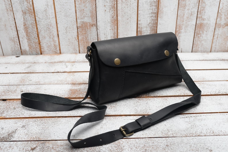 May include: A black leather crossbody bag with a flap closure and a long adjustable strap. The bag has a small pocket on the front and two brass snaps.