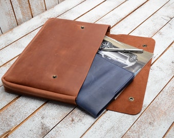 May include: A brown leather laptop sleeve with a blue leather notebook and a pen inside. The sleeve has a flap that closes with a snap.