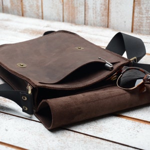 May include: A brown leather messenger bag with a black adjustable strap. The bag has a flap with a small pocket and a zipper closure. Sunglasses are tucked into the bag's open flap.