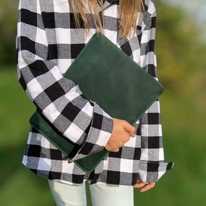 May include: A person wearing a black and white plaid shirt is holding a dark green leather clutch.