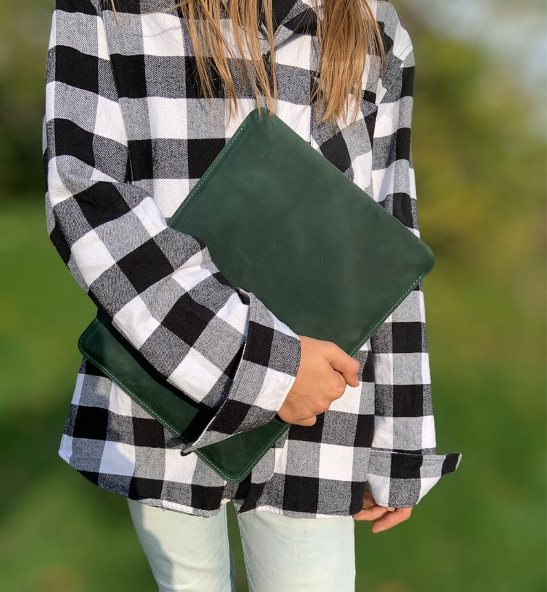 May include: A person wearing a black and white plaid shirt is holding a dark green leather clutch.