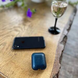 Puede incluir: Una funda de cuero azul oscuro para auriculares inal&aacute;mbricos se encuentra sobre una mesa de madera junto a un tel&eacute;fono inteligente negro y una copa de vino blanco.