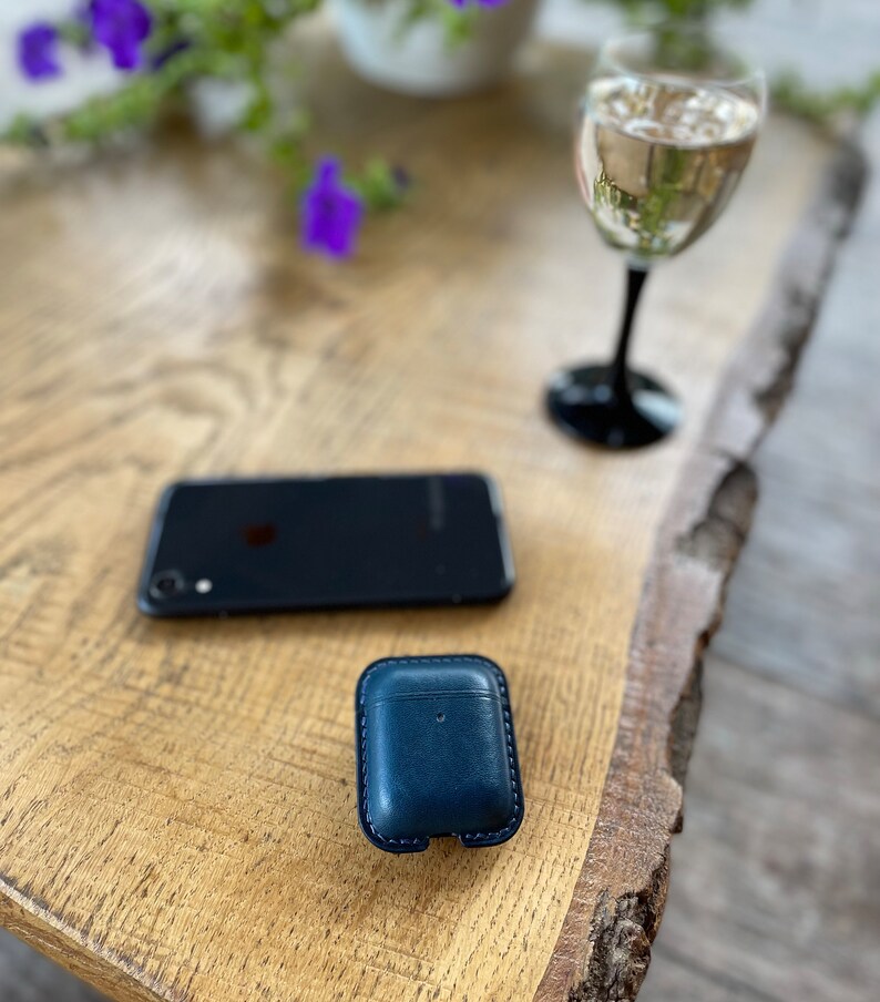 Puede incluir: Una funda de cuero azul oscuro para auriculares inal&aacute;mbricos se encuentra sobre una mesa de madera junto a un tel&eacute;fono inteligente negro y una copa de vino blanco.