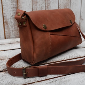 May include: A brown leather crossbody bag with a flap closure and a long adjustable strap. The bag has a small pocket on the front and a brass buckle on the strap.