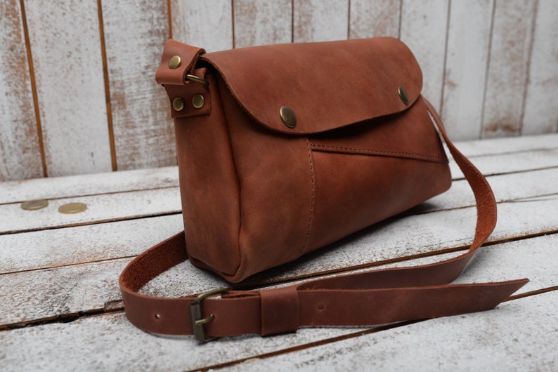 May include: A brown leather crossbody bag with a flap closure and a long adjustable strap. The bag has a small pocket on the front and a brass buckle on the strap.