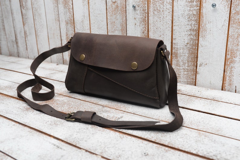 May include: A brown leather crossbody bag with a flap closure and a long adjustable strap. The bag has a distressed look and features two brass buttons on the flap.