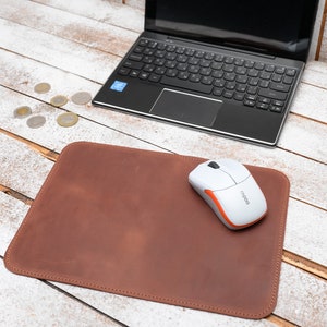 May include: A brown leather mouse pad with a stitched border, a white and orange wireless mouse, and a black laptop. Several coins are scattered nearby on a white wooden surface.