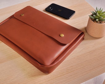 May include: A brown leather laptop sleeve with a flap closure and gold metal accents. The sleeve is on a light wood surface.