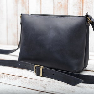 Peut inclure: Un sac &agrave; bandouli&egrave;re en cuir bleu fonc&eacute; avec une longue bandouli&egrave;re r&eacute;glable et une boucle dor&eacute;e. Le sac a une fermeture &agrave; glissi&egrave;re et une surface lisse et textur&eacute;e.