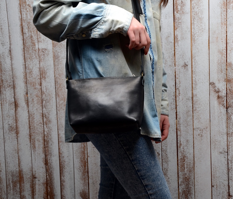 Pu&ograve; includere: Una borsa a tracolla in pelle nera con una lunga tracolla. La borsa &egrave; indossata da una persona che indossa una camicia di jeans e jeans.