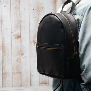 Pu&ograve; includere: Uno zaino in pelle nera con cerniere dorate e una forma arrotondata. Lo zaino &egrave; indossato da una persona.