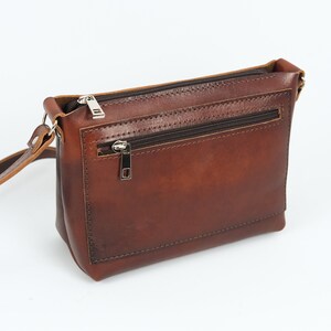 Pu&ograve; includere: Una borsa a tracolla in pelle marrone con una tasca con cerniera sul davanti. La borsa ha una lunga tracolla regolabile.