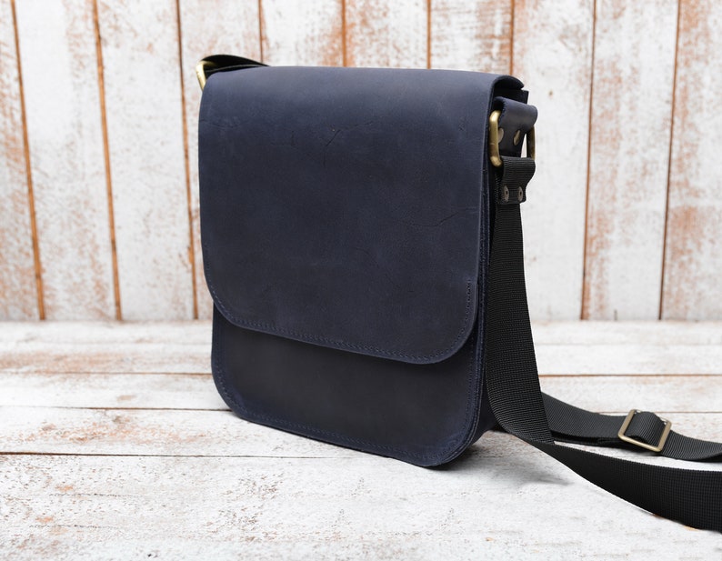 May include: A dark blue leather messenger bag with a black adjustable strap and brass hardware.