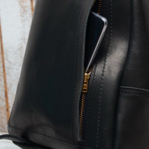 Pu&ograve; includere: Uno zaino in pelle nera con una tasca con cerniera dorata sul davanti. Uno smartphone &egrave; visibile all'interno della tasca.