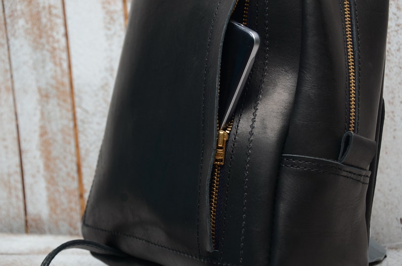 Pu&ograve; includere: Uno zaino in pelle nera con una tasca con cerniera dorata sul davanti. Uno smartphone &egrave; visibile all'interno della tasca.