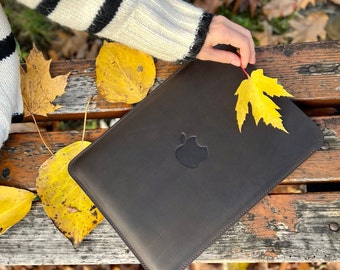 May include: A dark brown leather laptop sleeve with an Apple logo. The sleeve is lying on a wooden surface with autumn leaves scattered around it.