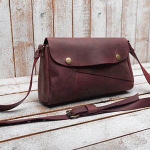 May include: A burgundy leather crossbody bag with a flap closure and a long adjustable strap. The bag has a distressed look and features a contrasting brown leather patch on the front.