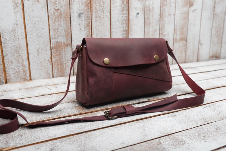 May include: A burgundy leather crossbody bag with a flap closure and a long adjustable strap. The bag has a distressed look and features a contrasting brown leather patch on the front.