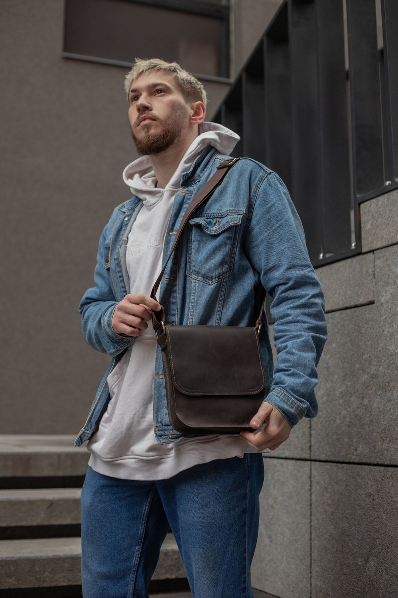May include: A man wearing a white hoodie and a blue denim jacket is carrying a brown leather messenger bag.