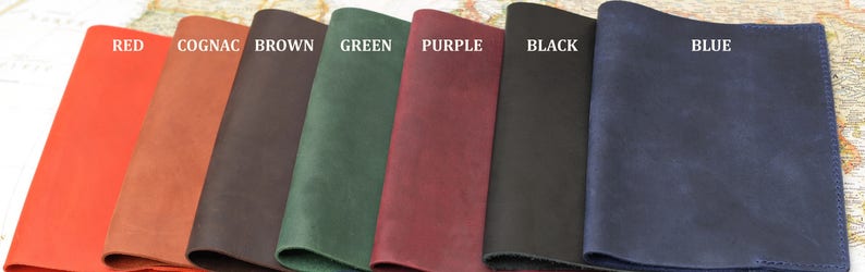 May include: Seven leather notebook covers in different colors: red, cognac, brown, green, purple, black, and blue. The covers are all made of the same type of leather and have a smooth, textured finish.