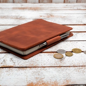 May include: A brown leather bound journal with a pen tucked into the elastic band. The journal is lying on a white wooden surface with several coins scattered around it.