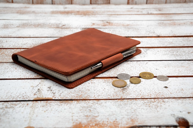 May include: A brown leather bound journal with a pen tucked into the elastic band. The journal is lying on a white wooden surface with several coins scattered around it.