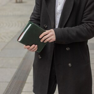 May include: A person wearing a black coat and white shirt is holding a dark green leather bound journal.