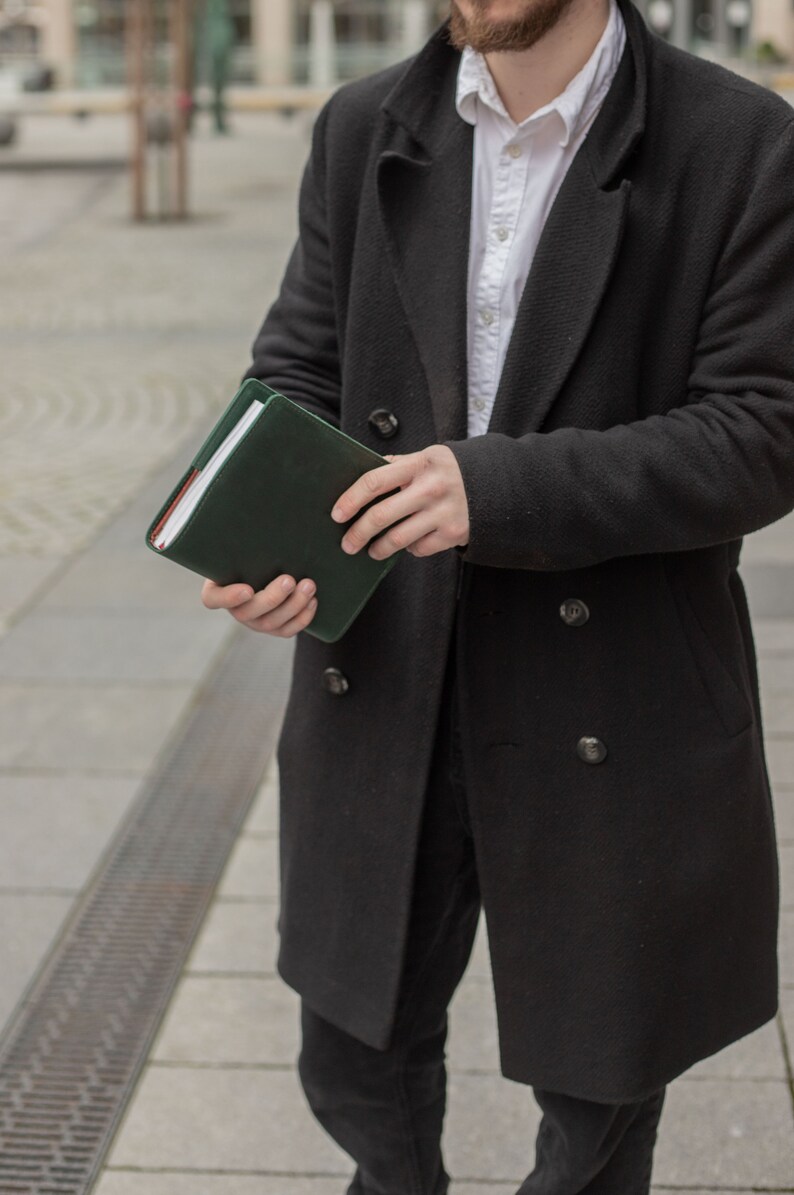 May include: A person wearing a black coat and white shirt is holding a dark green leather bound journal.