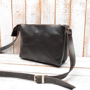 May include: A brown leather crossbody bag with a zipper closure and a brown strap. The bag has a stitched detail around the edges.