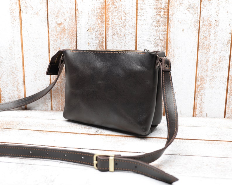 May include: A brown leather crossbody bag with a zipper closure and a brown strap. The bag has a stitched detail around the edges.