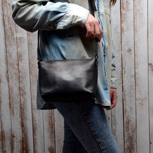 May include: A black leather crossbody bag with a long strap. The bag is worn by a person wearing a denim shirt and jeans.