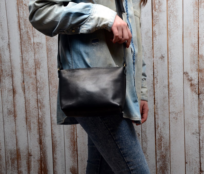 May include: A black leather crossbody bag with a long strap. The bag is worn by a person wearing a denim shirt and jeans.