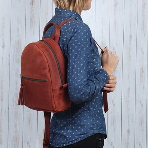 Pu&ograve; includere: Uno zaino in pelle marrone con un fiocco e una tasca con cerniera. Lo zaino &egrave; indossato da una persona che indossa una camicia a pois blu e bianchi.