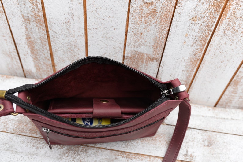 Peut inclure: Un sac &agrave; bandouli&egrave;re en cuir bordeaux avec une fermeture &agrave; glissi&egrave;re et un tirette de fermeture &eacute;clair noire. Le sac a une petite poche int&eacute;rieure et une longue bandouli&egrave;re.