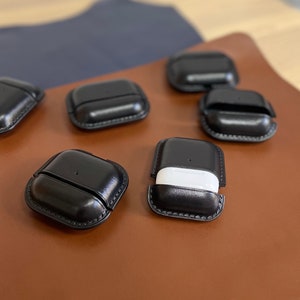 Puede incluir: Seis estuches de cuero negro para Airpods, con un estuche abierto para revelar un estuche Airpod blanco en el interior.