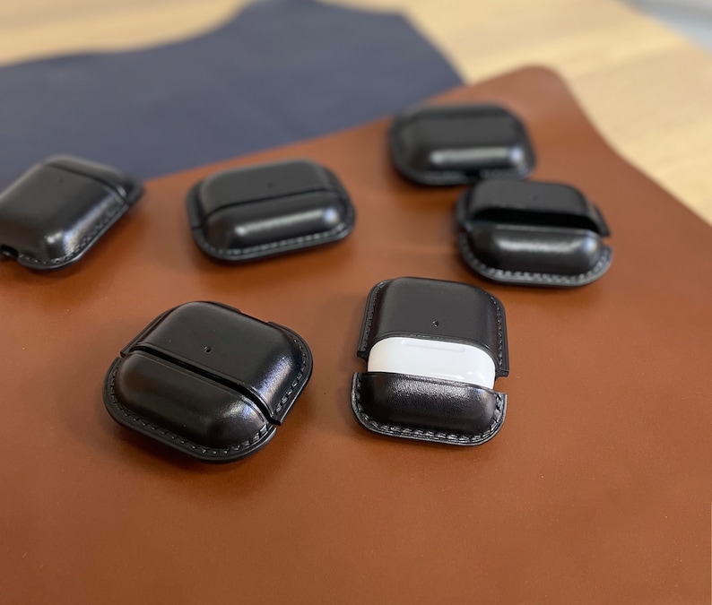 Puede incluir: Seis estuches de cuero negro para Airpods, con un estuche abierto para revelar un estuche Airpod blanco en el interior.