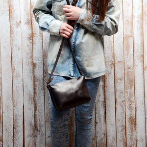 May include: A brown leather crossbody bag with a long adjustable strap. The bag is worn by a person wearing a light blue denim shirt and jeans.