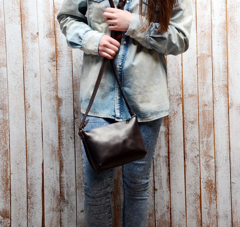 May include: A brown leather crossbody bag with a long adjustable strap. The bag is worn by a person wearing a light blue denim shirt and jeans.