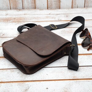 May include: A brown leather messenger bag with a black adjustable strap and a flap closure. The bag is lying on a white wooden surface with a pair of brown sunglasses next to it.