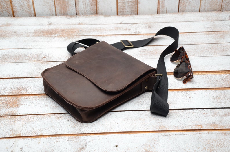 May include: A brown leather messenger bag with a black adjustable strap and a flap closure. The bag is lying on a white wooden surface with a pair of brown sunglasses next to it.