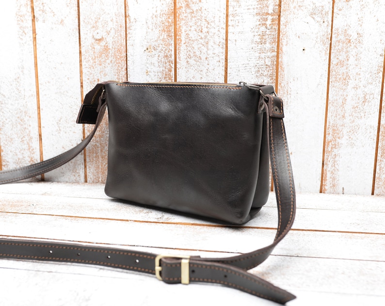 Pu&ograve; includere: Una borsa a tracolla in pelle marrone con chiusura a zip e una tracolla marrone. La borsa ha un design cucito attorno ai bordi.
