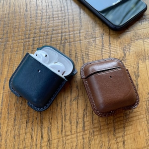 Puede incluir: Dos estuches de cuero para auriculares inal&aacute;mbricos. Un estuche es azul oscuro y el otro es marr&oacute;n. Ambos estuches tienen un interior blanco.