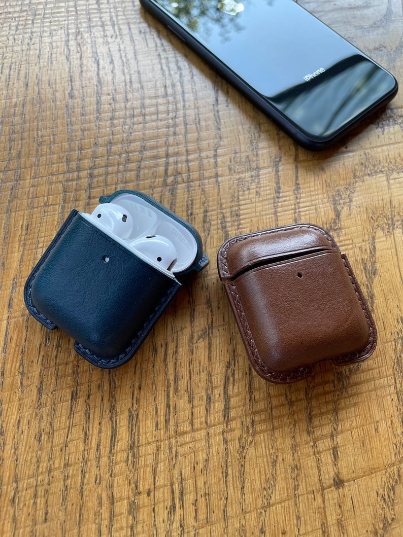 Puede incluir: Dos estuches de cuero para auriculares inal&aacute;mbricos. Un estuche es azul oscuro y el otro es marr&oacute;n. Ambos estuches tienen un interior blanco.