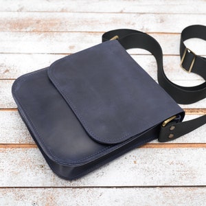 May include: A dark blue leather messenger bag with a black adjustable strap and brass hardware.