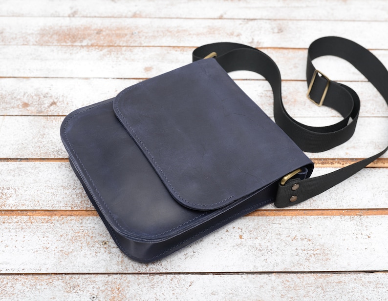 May include: A dark blue leather messenger bag with a black adjustable strap and brass hardware.